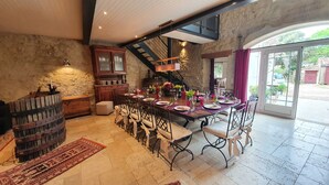 A spacious dining area with a long wooden table set for a meal, a rustic stone wall, and a view through a glass door to the outside.