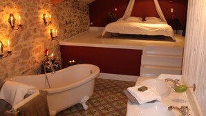 A bedroom with a stone wall, a white bed, a canopy, a bathtub, and a bathroom with a sink and mirror.