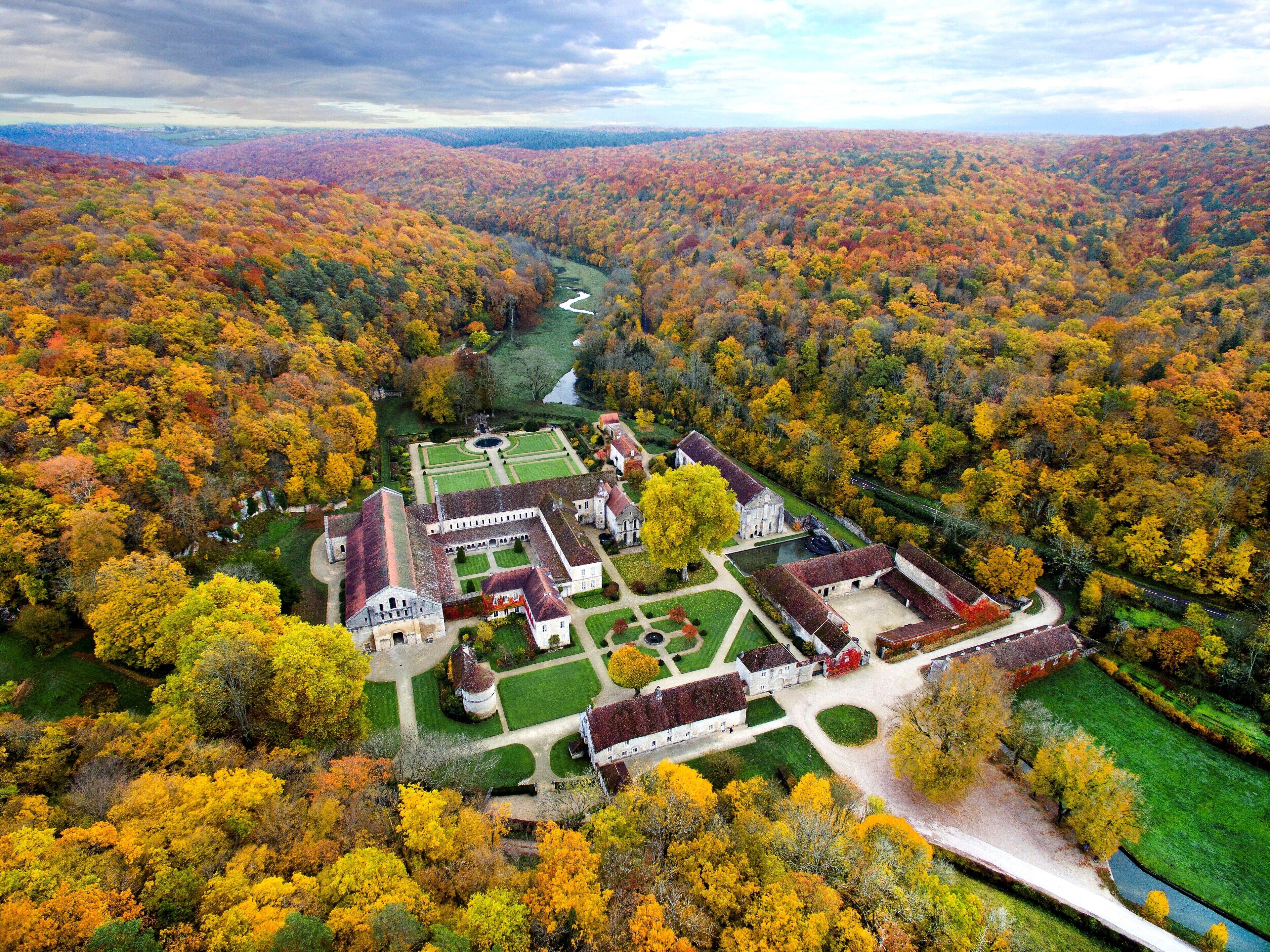 An aerial view of a historic estate with a formal garden, a river, and a dense forest in autumn colors.