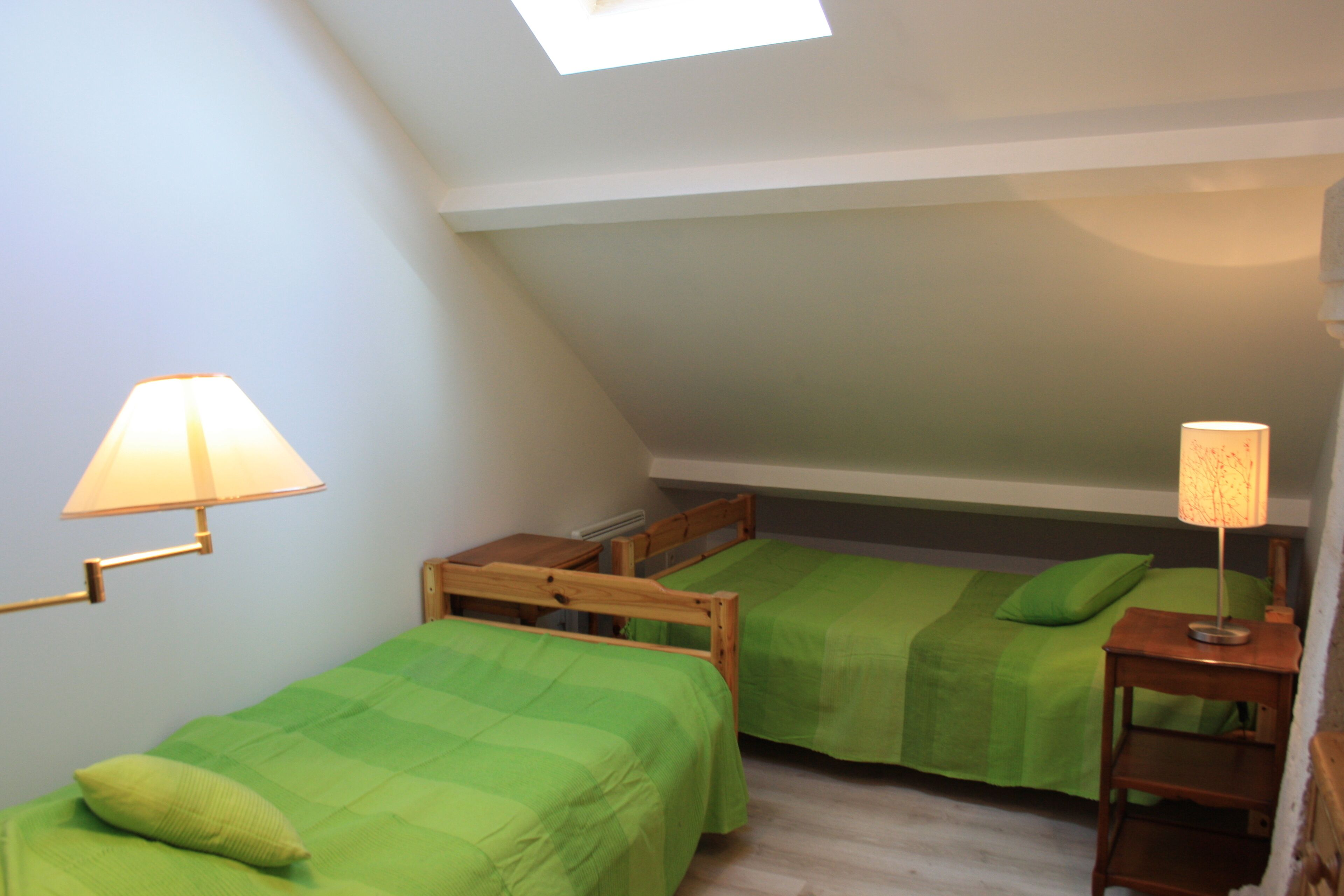 A small, cozy room with two wooden beds, each with a green bedspread and a pillow. There are two bedside tables with lamps, and the room has a sloped ceiling with a skylight.