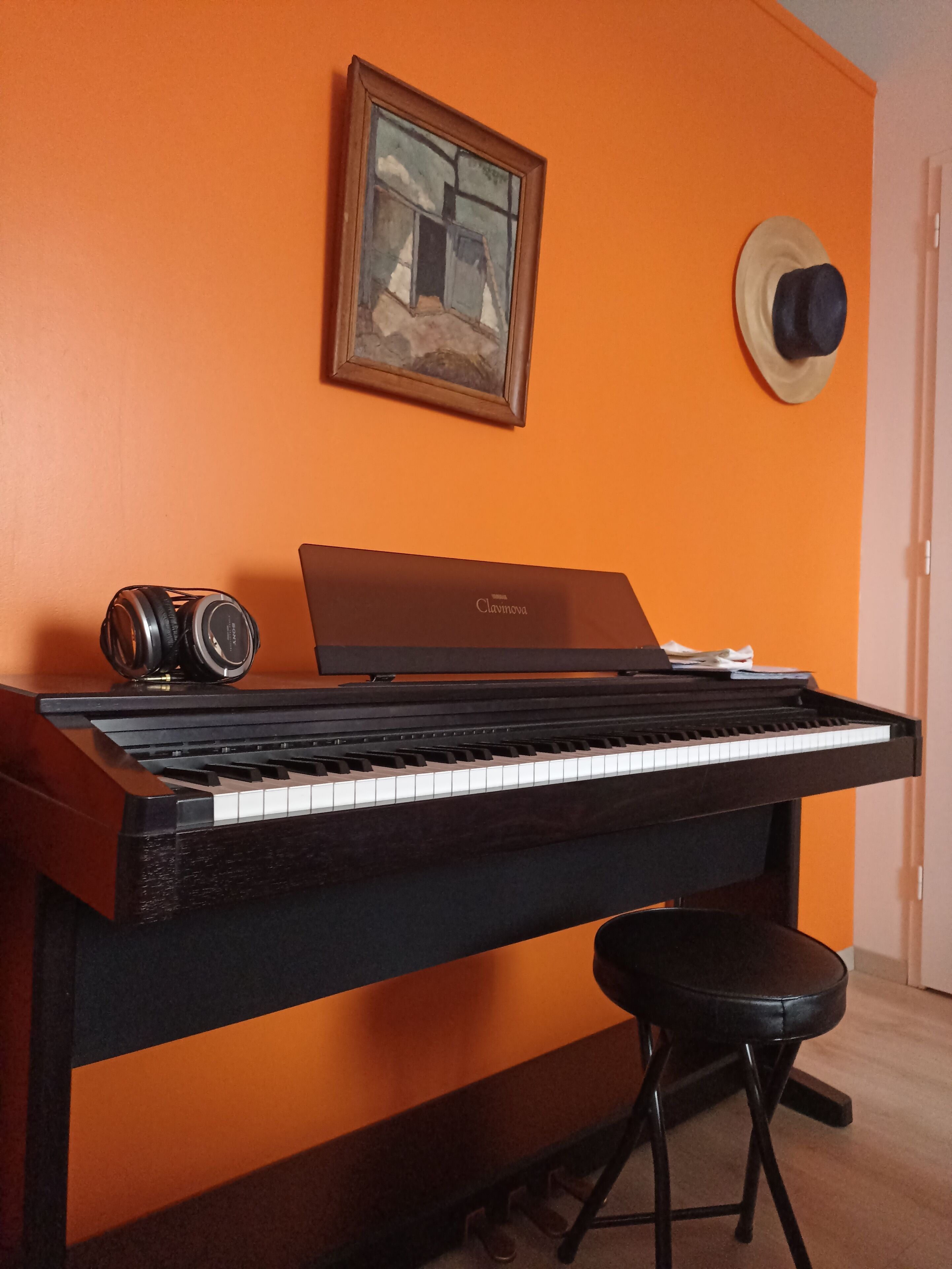 Ett piano med hörlurar och en bärbar dator ovanpå, en pall och en hatt som hänger på väggen.