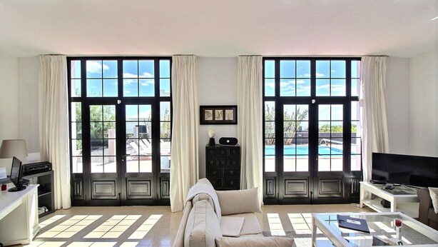 A spacious living room with a sofa, coffee table, and TV, large windows with curtains, and a pool view. - Luxury villa with private heated pool, WIFI, views of the sea and volcanoes (TIAS)