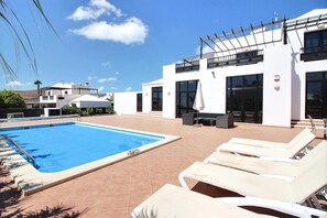A modern outdoor pool area with a rectangular pool, white lounge chairs, and a white building with large windows and balconies.