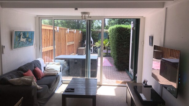 A living room with a sofa, a coffee table, a TV, and a sliding glass door leading to a patio with a wooden fence and greenery. - A cozy little nest in Biscarosse Lac, Superb high-end full option duplex (Biscarrosse)