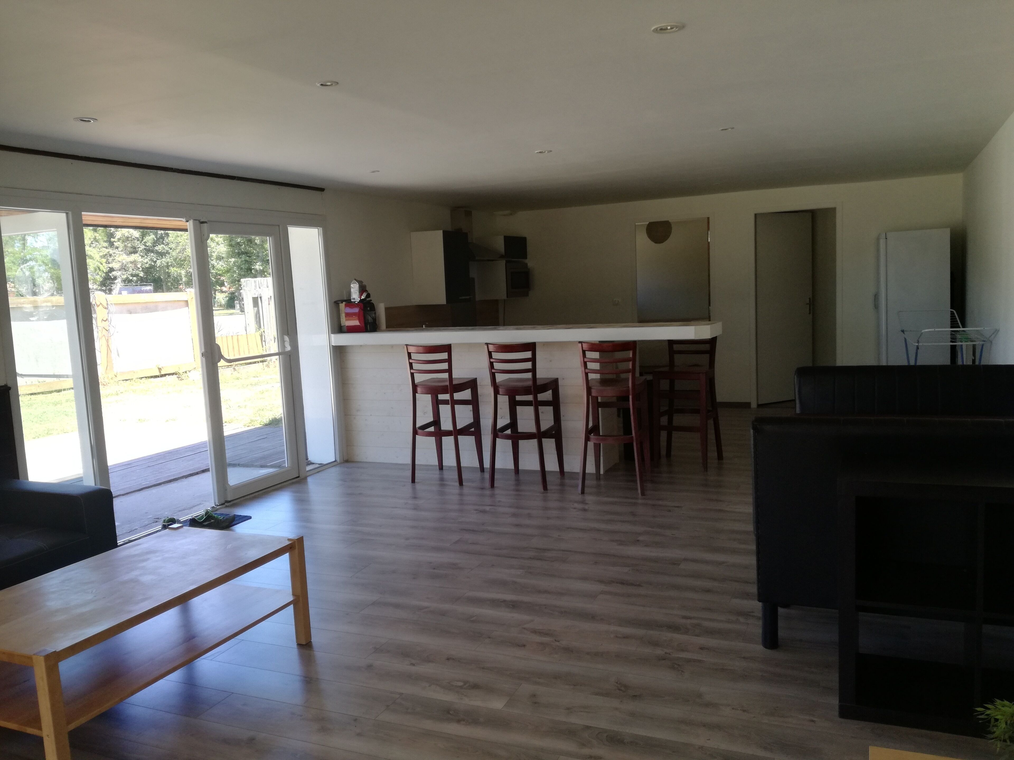 Un salón amplio con suelo de madera, un sofá negro, una mesa baja de madera y una zona de cocina con mostrador y tres sillas de madera.