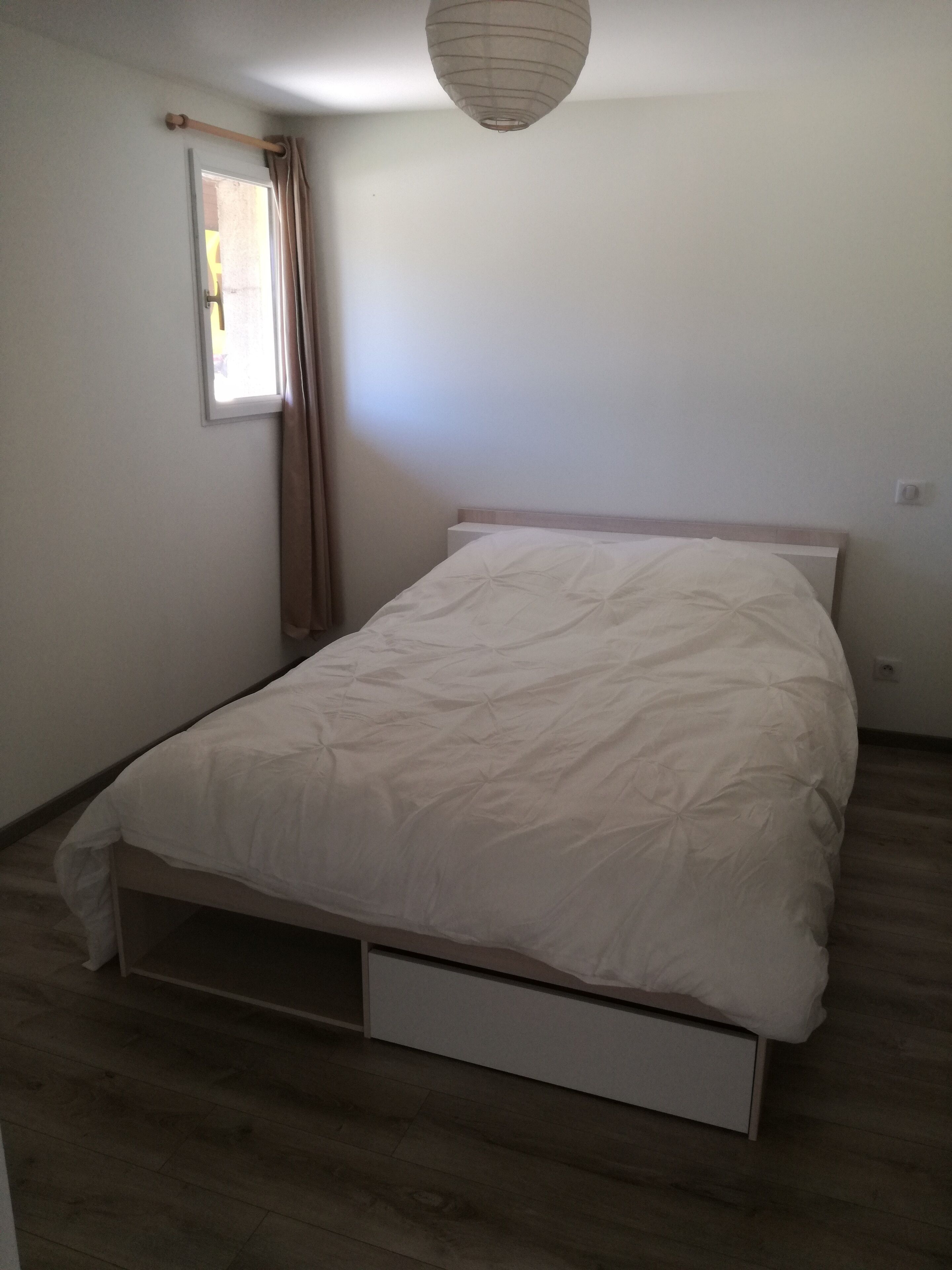 A bedroom with a single bed, a window with curtains, a bedside table, and a hanging light fixture.