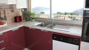 A modern kitchen with red cabinets, a stainless steel sink, and a dishwasher. There are two potted plants on the counter and a red toaster. -  Apartment with Exceptional Panorama (Villefranche sur Mer)