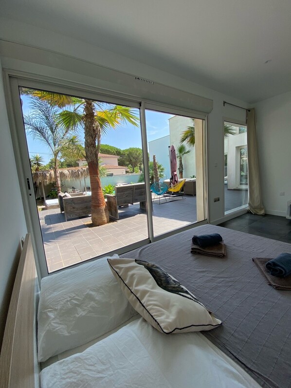 A bedroom with a large sliding glass door leading to an outdoor area with a pool, palm trees, and a chair. - Your Exceptional Getaway in Le Grau-d'Agde: Architect-Designed Villa with Private Pool (Agde)