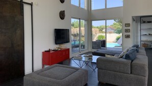 A modern living room with a large TV, grey sofas, a red cabinet, and a sliding glass door leading to a pool area. - Your Exceptional Getaway in Le Grau-d'Agde: Architect-Designed Villa with Private Pool (Agde)