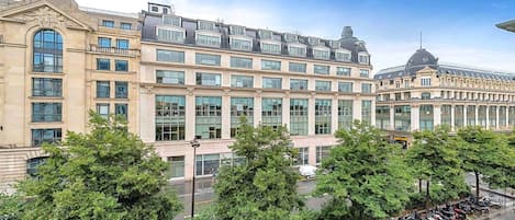 Un grand immeuble à étages, de style architectural classique, doté d’une cour intérieure arborée et d’une rangée de motos garées.