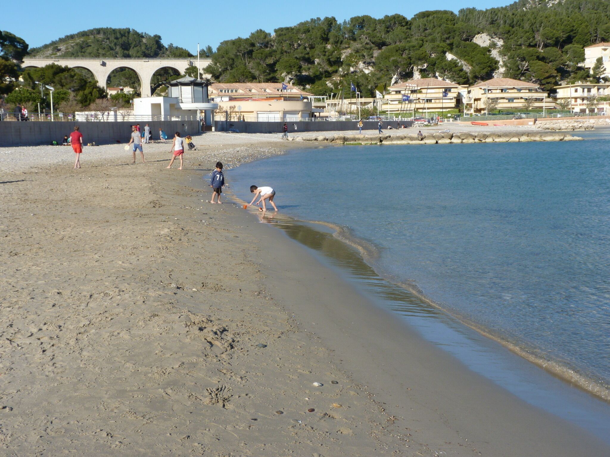 Una playa con gente caminando y jugando cerca del agua, un puente de piedra y edificios al fondo.