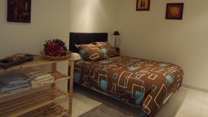 Un dormitorio con una cama, una mesita de noche con una lámpara, un estante con toallas dobladas y dos cuadros en la pared.