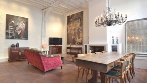 A spacious living room with a large wooden dining table, a red sofa, and a fireplace. There are paintings on the walls and a chandelier above the table.