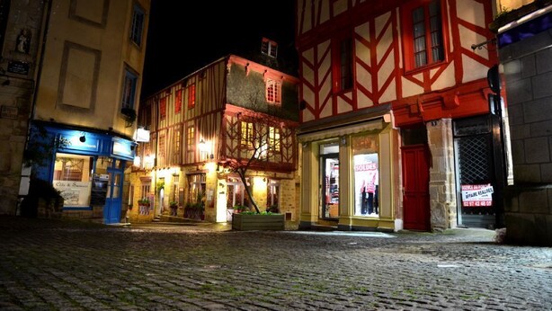 A cobblestone street at night with traditional half-timbered buildings, a café, and a shop with a sale sign. - Bright and comfortable house in a quiet area in a wooded park near the beaches. (Plouharnel)