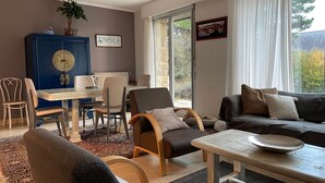 A cozy living room with a sofa, chairs, a dining table, and a large window with curtains. - Bright and comfortable house in a quiet area in a wooded park near the beaches. (Plouharnel)