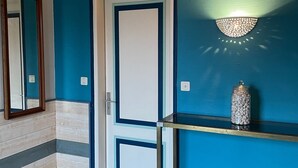 A hallway with a blue wall, a white door, a mirror, and a wall-mounted light fixture. - Bright and comfortable house in a quiet area in a wooded park near the beaches. (Plouharnel)