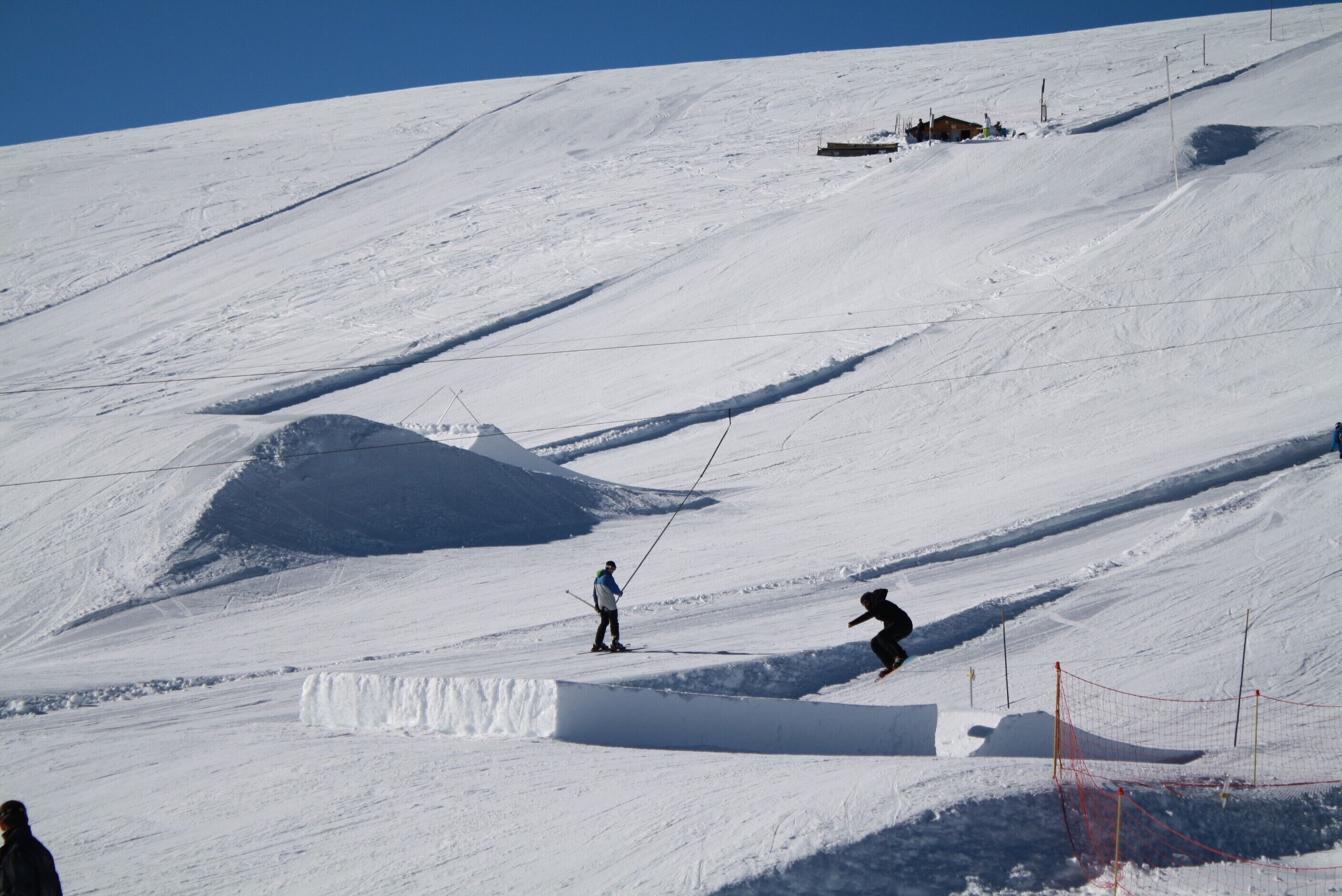 A snowy ski slope with a person skiing down a slope and another standing on a flat area.