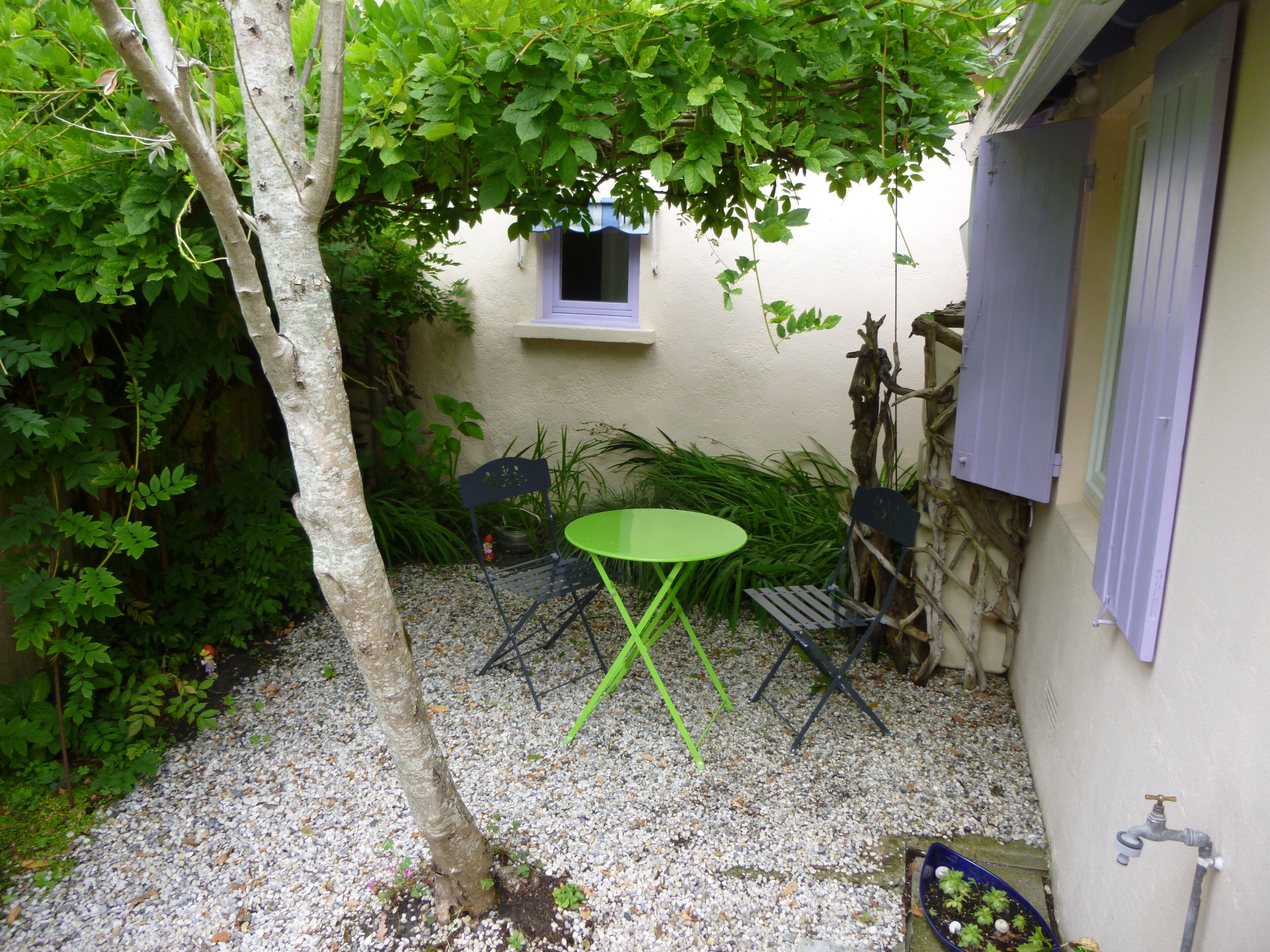 Eine kleine Terrasse im Freien mit Tisch und Stühlen, umgeben von Pflanzen und einer weißen Mauer.