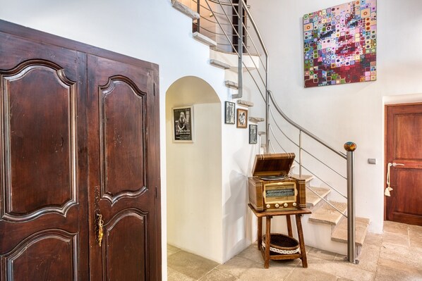 Une porte en bois, un escalier avec une rampe en métal, une radio ancienne posée sur une petite table et un tableau abstrait aux couleurs vives accroché au mur.