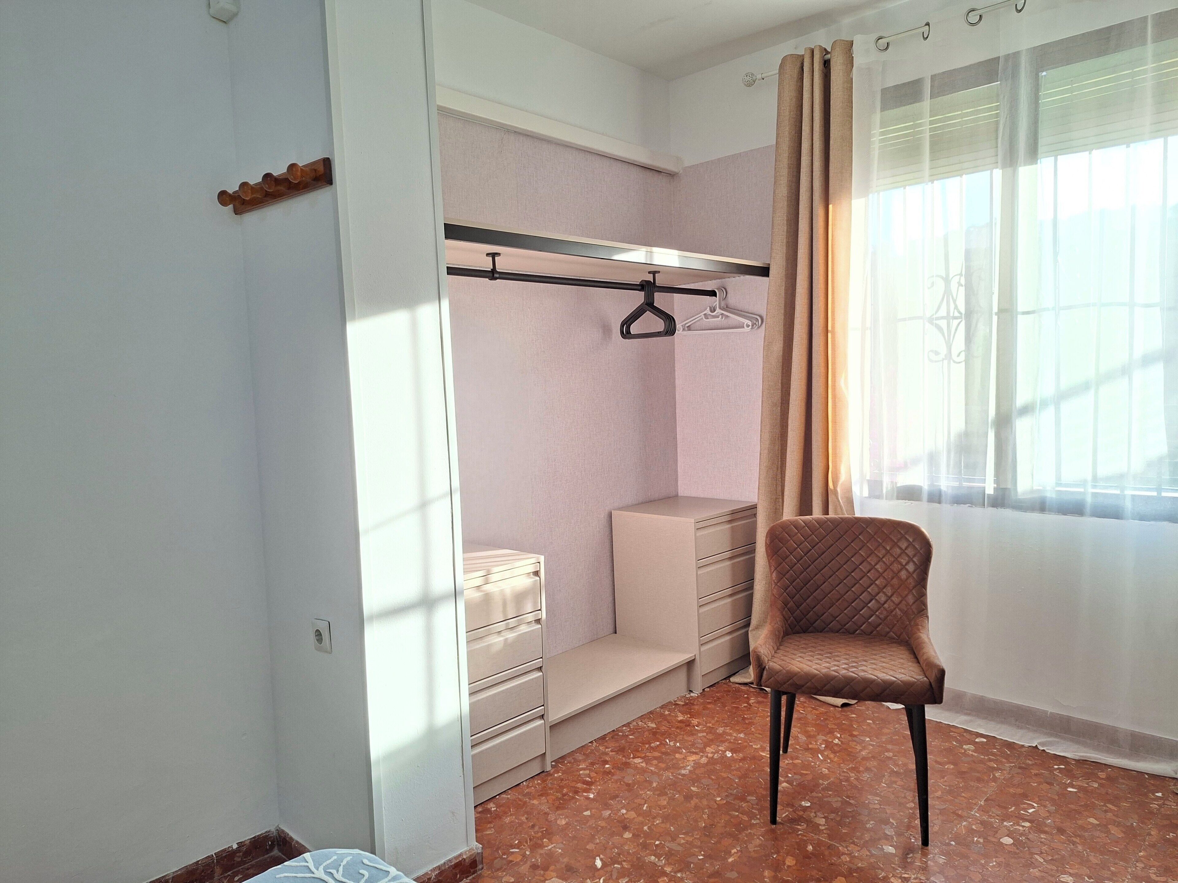 Ein Schlafzimmer mit Bett, Sessel, Kleiderschrank und einem fenster mit Vorhängen.