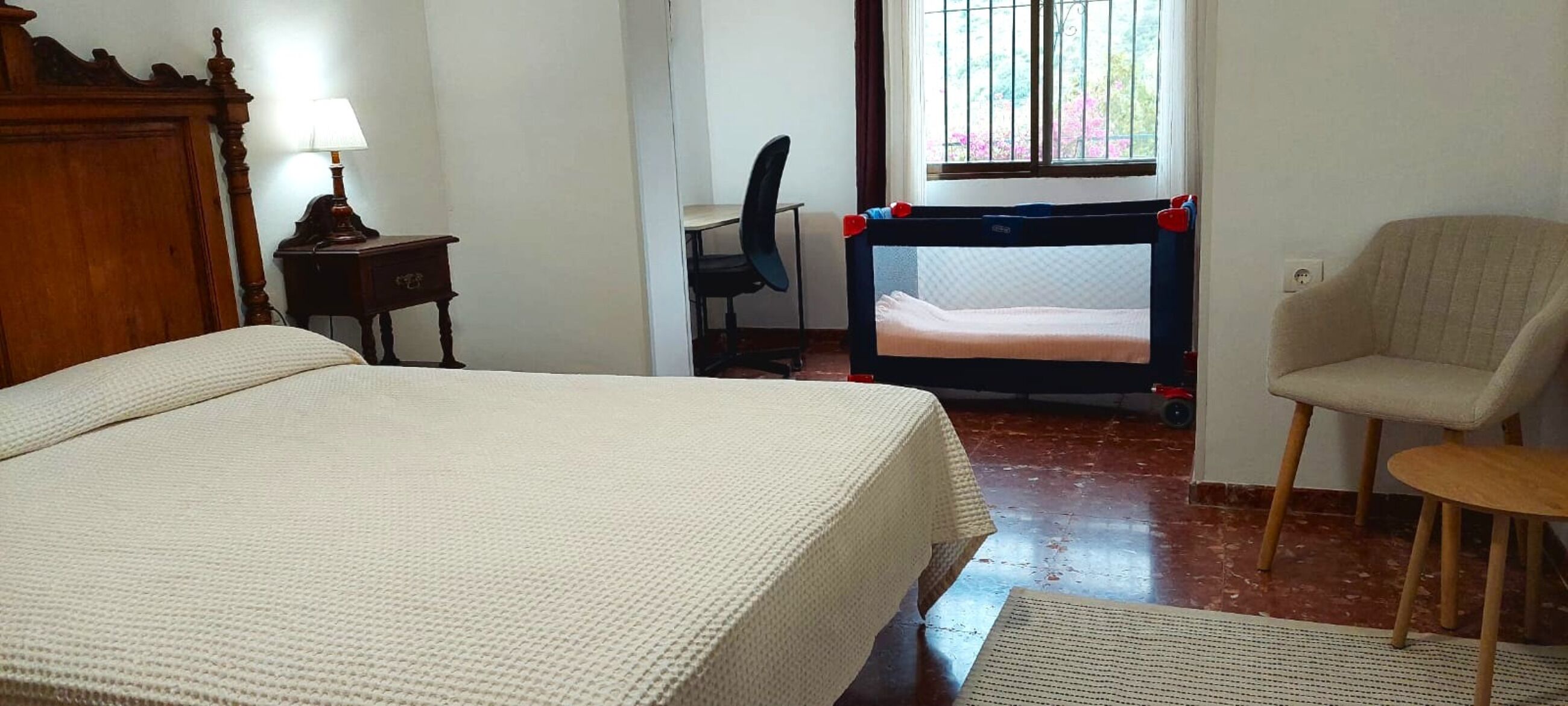 A bedroom with a bed, a chair, a desk, a window with bars, and a view of greenery outside.