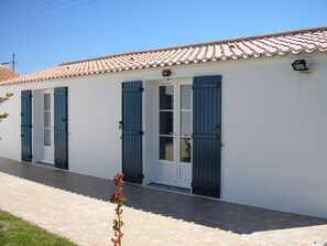 A white building with a tiled roof, two doors with blue shutters, and a small flower in the foreground.
