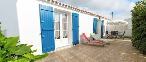Een wit gebouw met blauwe luiken, een betegeld terras en een tafel met stoelen.