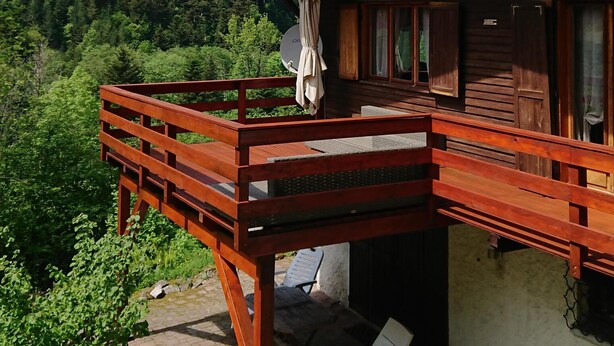 Eine Holzhütte mit Balkon, weißen Lounge-Sesseln und Blick auf einen bewaldeten Berg.