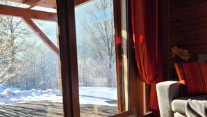 Ein Zimmer mit einer großen Schiebeglasdoor zum verschneiten Außenbereich, einem grauen Sofa, einem roten Vorhang und einer Holzbalkendecke.