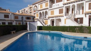 A swimming pool with a clear blue water surface, surrounded by a white building with balconies and a tiled roof. - Cozy bungalow, 2 min walk from the beach. Wi-Fi (Torrevieja)
