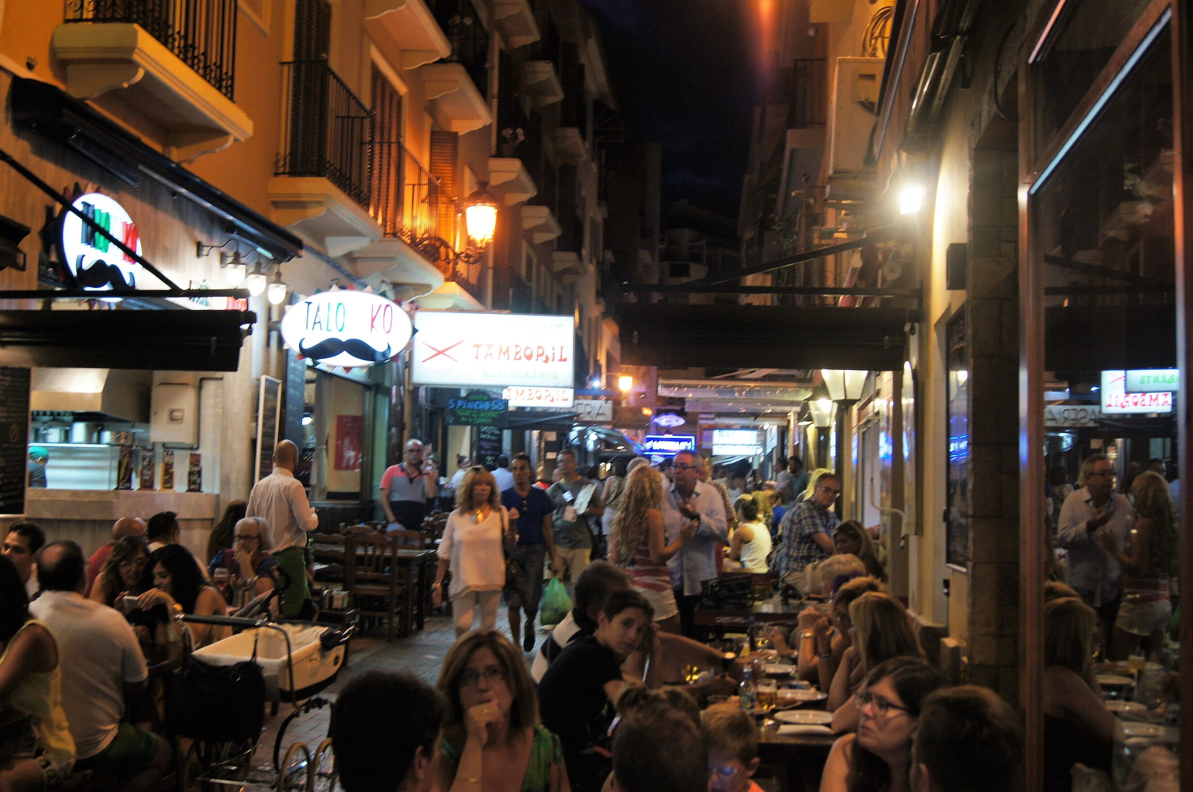 A crowded street scene at night with people walking and dining at outdoor cafes.