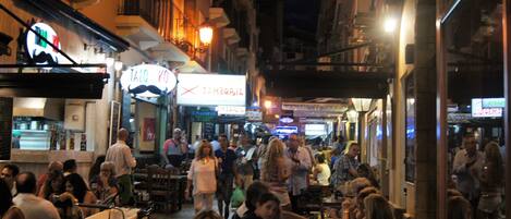 A crowded street scene at night with people walking and dining at outdoor cafes.