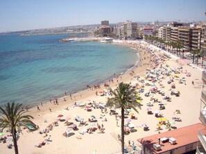 Una spiaggia con molti ombrelloni e persone, un mare cristallino e edifici sullo sfondo.