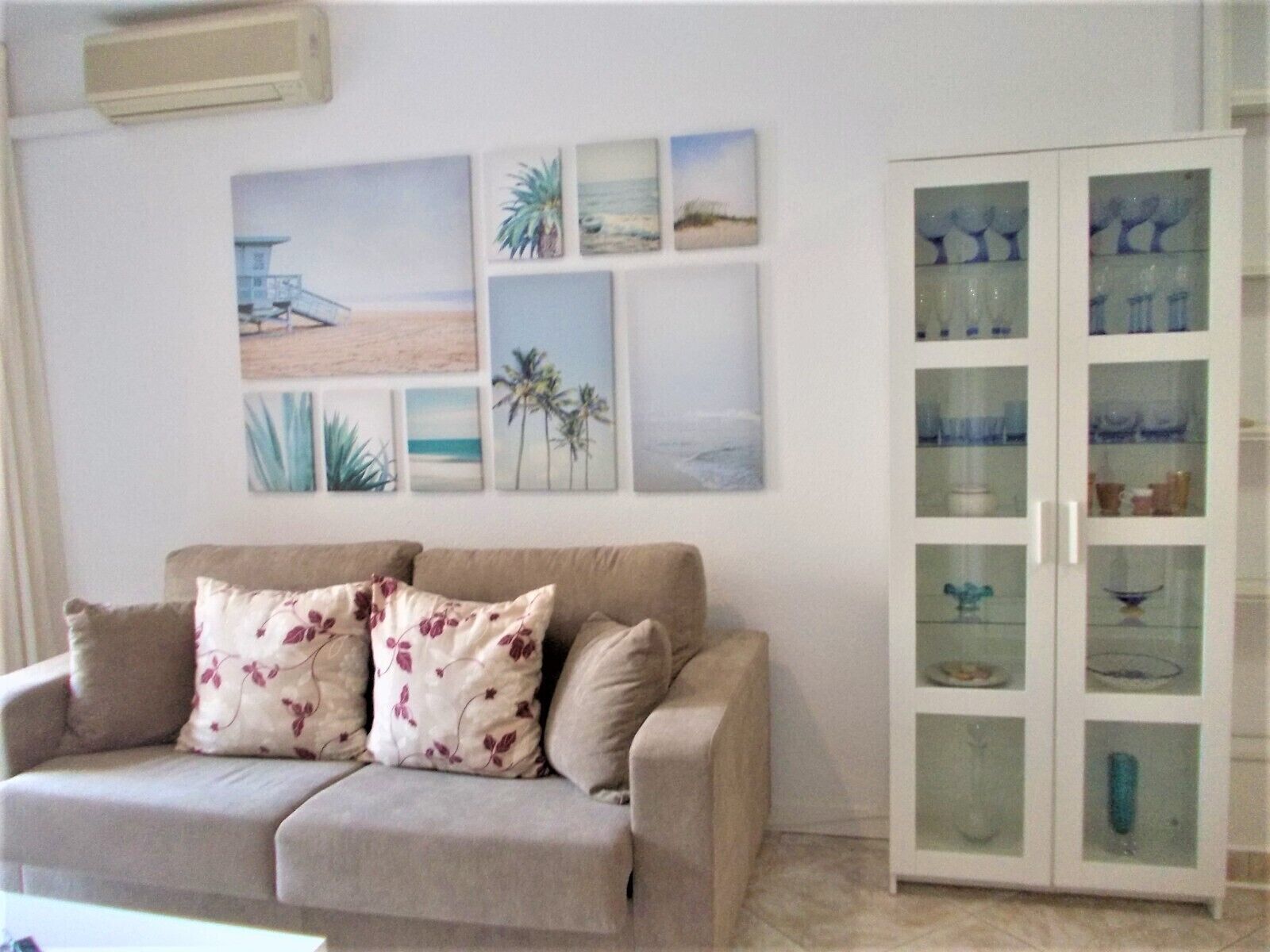 Een woonkamer met een beige bank, bloemenkussens en een glazen kast met decoratieve spullen.