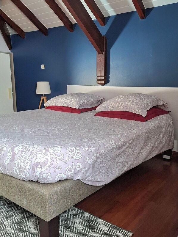 A bedroom with a large bed, blue walls, wooden beams, a bedside table with a lamp, and patterned bedding. - SEASONAL RENTAL VILLA STANDING 3/4 PERS (2 ADULT / 2 NFE). POOL SPA SAUNA (Le Moule)