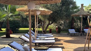 A series of sun loungers with white cushions, arranged in rows, under thatched umbrellas, set on a paved patio with a lush garden in the background. - Marrakech Villa for 6-16 people - Exclusive Offer - 450 sq.m and 16 x 5.5 m pool (Marrakech)