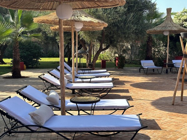 A series of sun loungers with white cushions, arranged in rows, under thatched umbrellas, set on a paved patio with a lush garden in the background.