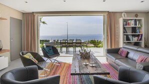 Een moderne woonkamer met een grote schuifpui naar een balkon met uitzicht op zee. Er is een bank, een salontafel en een boekenkast.
