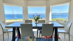 Een eetruimte met een tafel en stoelen, een vaas met bloemen en grote ramen met uitzicht op zee.