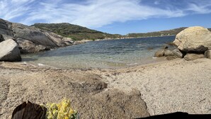 A rocky beach with clear water, large boulders, and a distant view of hills.