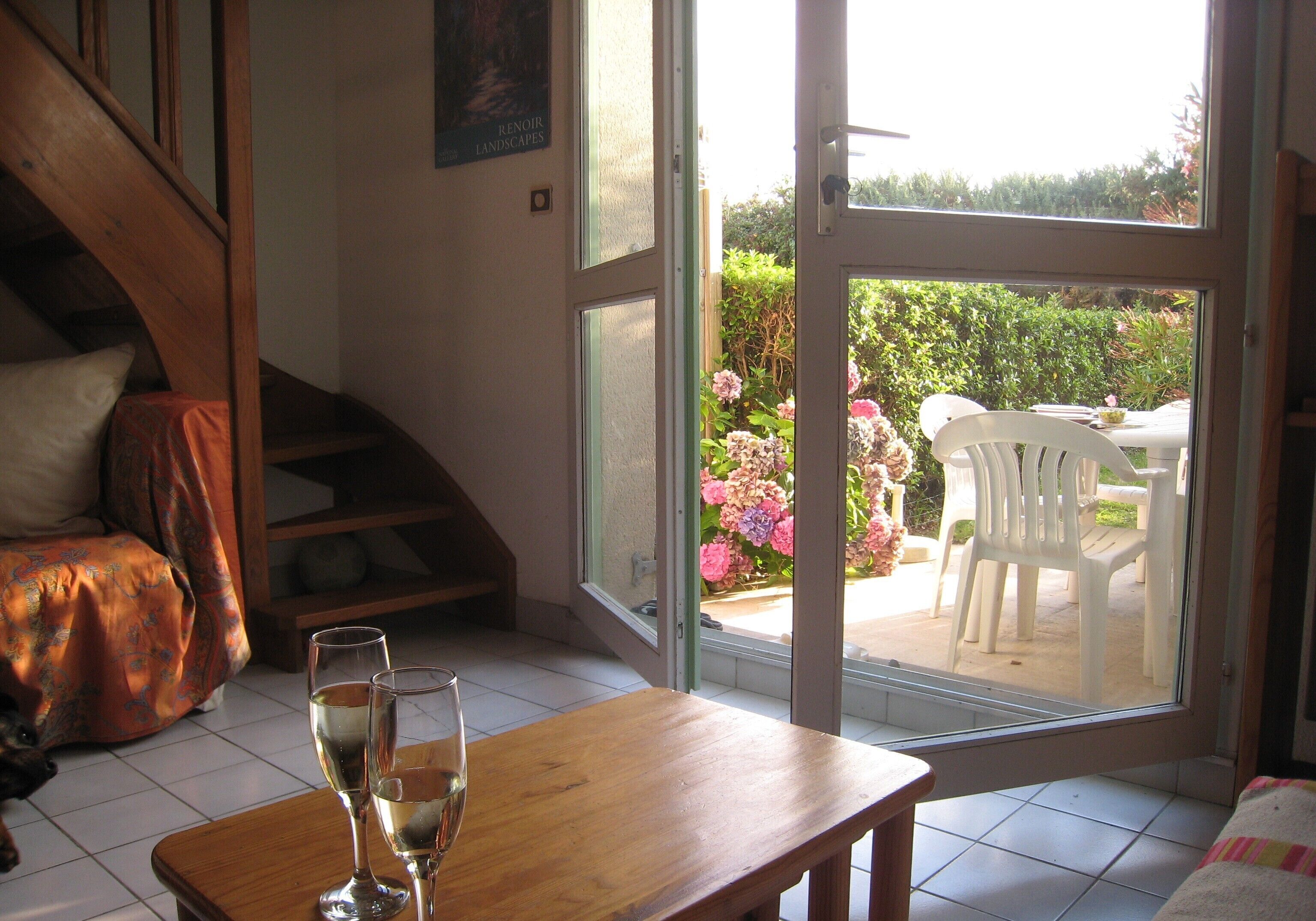 Une pièce avec une table en bois, deux verres de vin blanc, un canapé et une vue sur un jardin, visible à travers une porte vitrée.
