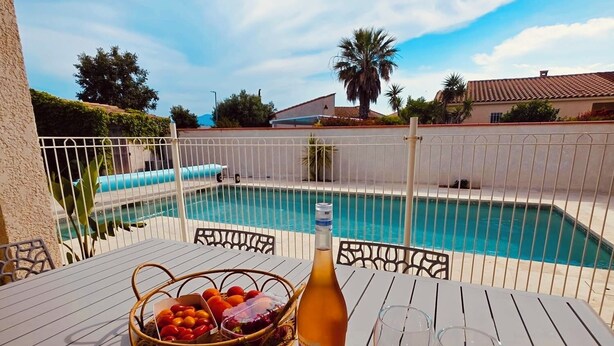 Une table placée au bord de la piscine, avec une bouteille et des verres, un panier de fruits et une vue sur la piscine.