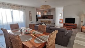 Un salon moderne comprenant un coin repas, un canapé gris, des fauteuils en osier et un téléviseur.