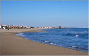 En strand med klart skys og sandet bred, samt bygninger i baggrunden.