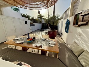 An outdoor dining area with a wooden table set for six, surrounded by white walls and a large potted plant.