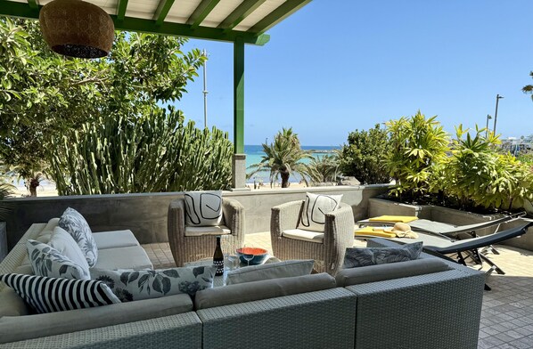 A covered outdoor seating area with wicker furniture, a bottle, and a bowl on a table, overlooking a beach.