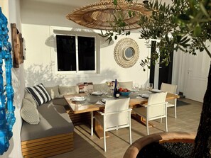 A dining area with a wooden table set for four, a sofa with striped cushions, a large mirror, and a wicker umbrella.