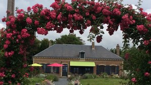 Un edificio in mattoni con una tenda verde, circondato da un giardino con fiori rosa e un vialetto sterrato che conduce all'ingresso.