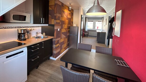 Cocina pequeña con microondas, cafetera y lavabo. Hay una mesa de comedor con sillas y una pared roja.