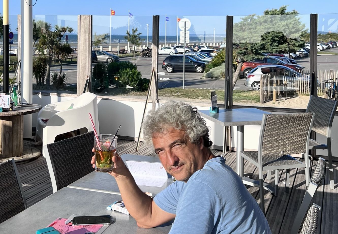 Ein Mann sitzt an einem Tisch im Freien, vor ihm ein Getränk, eine Speisekarte und ein Smartphone.